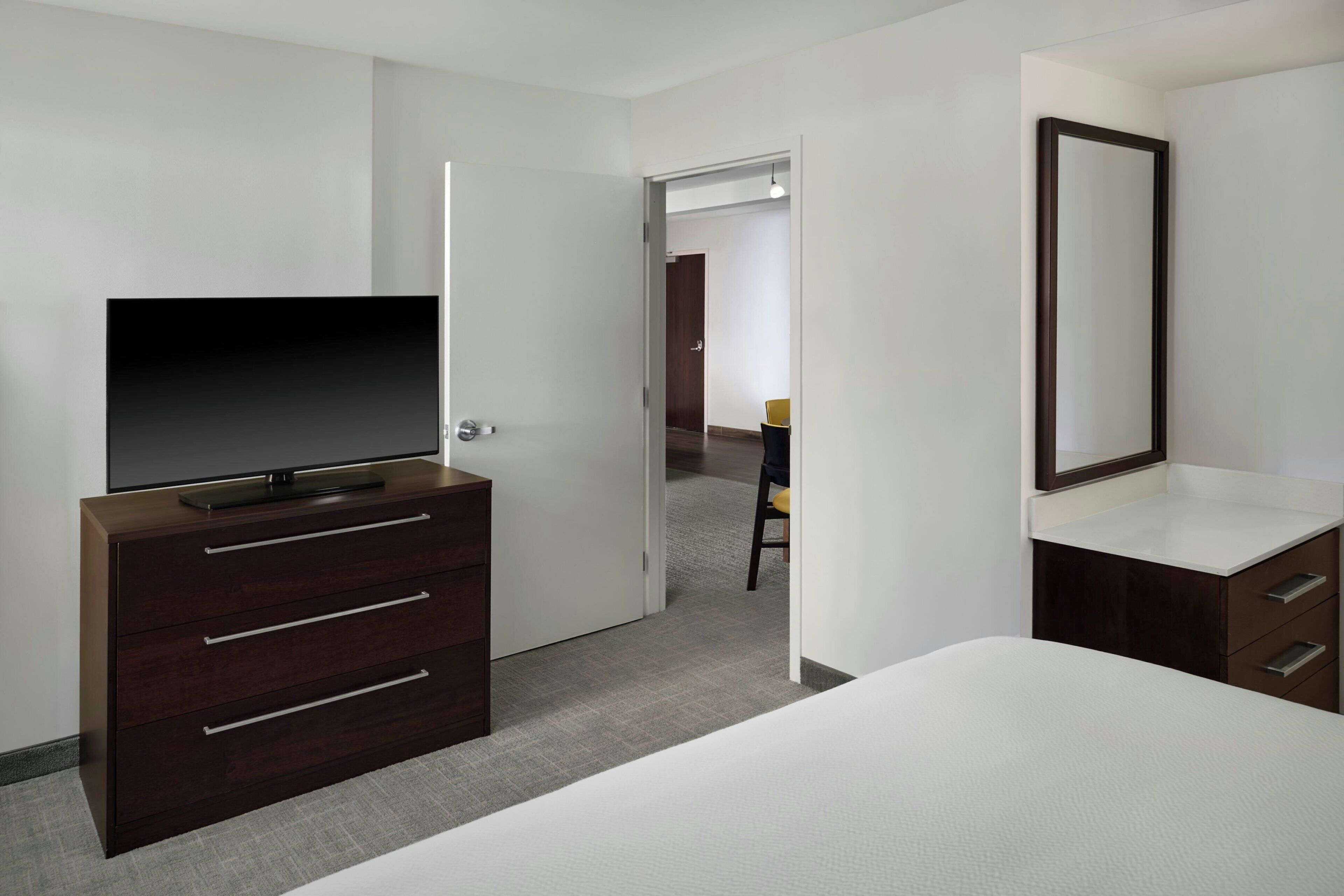 A hotel room with a bed, a dresser with drawers, a television, a mirror, and a door leading to another room.
