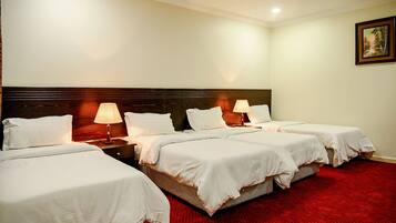 A hotel room with four single beds, each with white bedding and a wooden headboard. There are two bedside lamps on each side of the beds. The room has a red carpet and a framed picture on the wall.