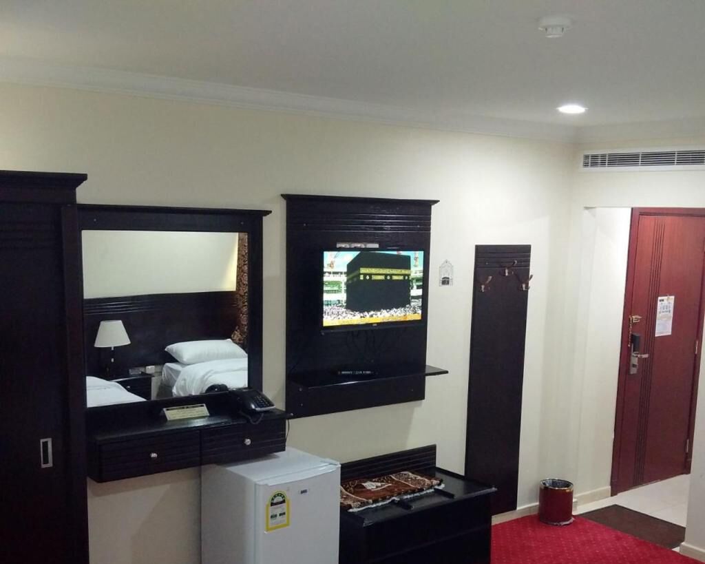 A hotel room with a bed, a TV mounted on the wall, a small refrigerator, and a red carpeted area.