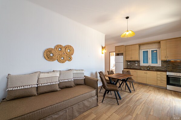 A modern kitchen with wooden cabinets, a dining table with chairs, and a sofa with patterned cushions. - Manolia View Mykonos (Mykonos)