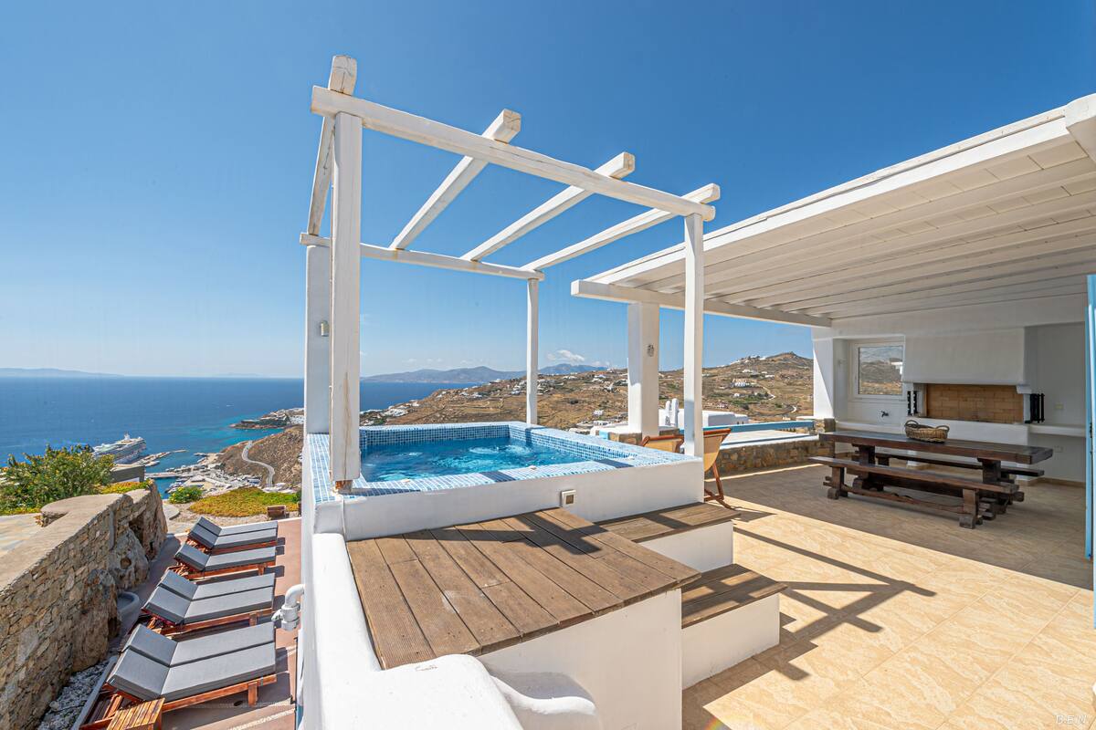 Una terraza en la azotea con jacuzzi, sillas de descanso y una cubierta de madera con vistas panorámicas al mar y las colinas.