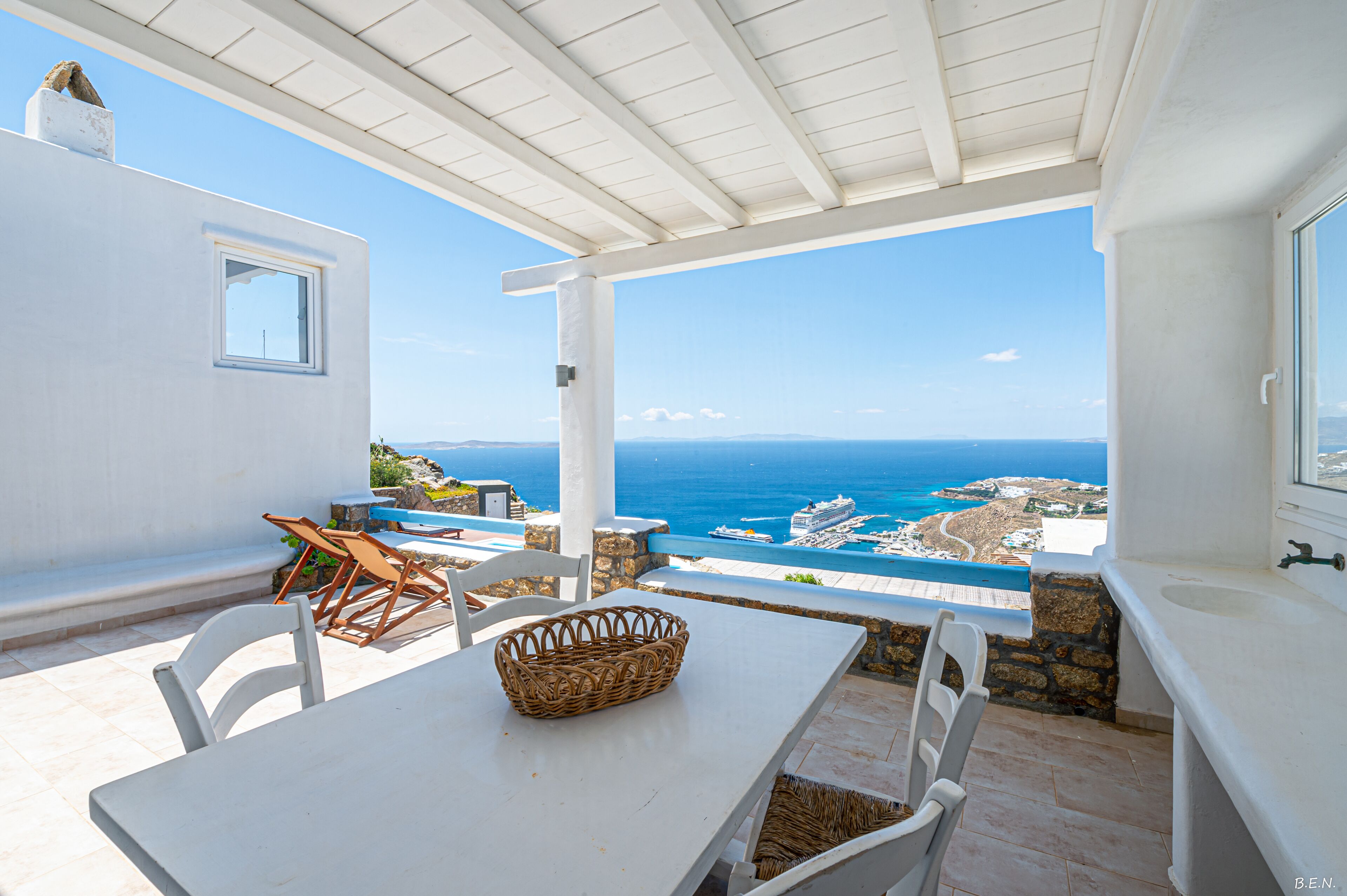 A terrace with a white table and chairs, a wicker basket, and a view of the sea and coastline.