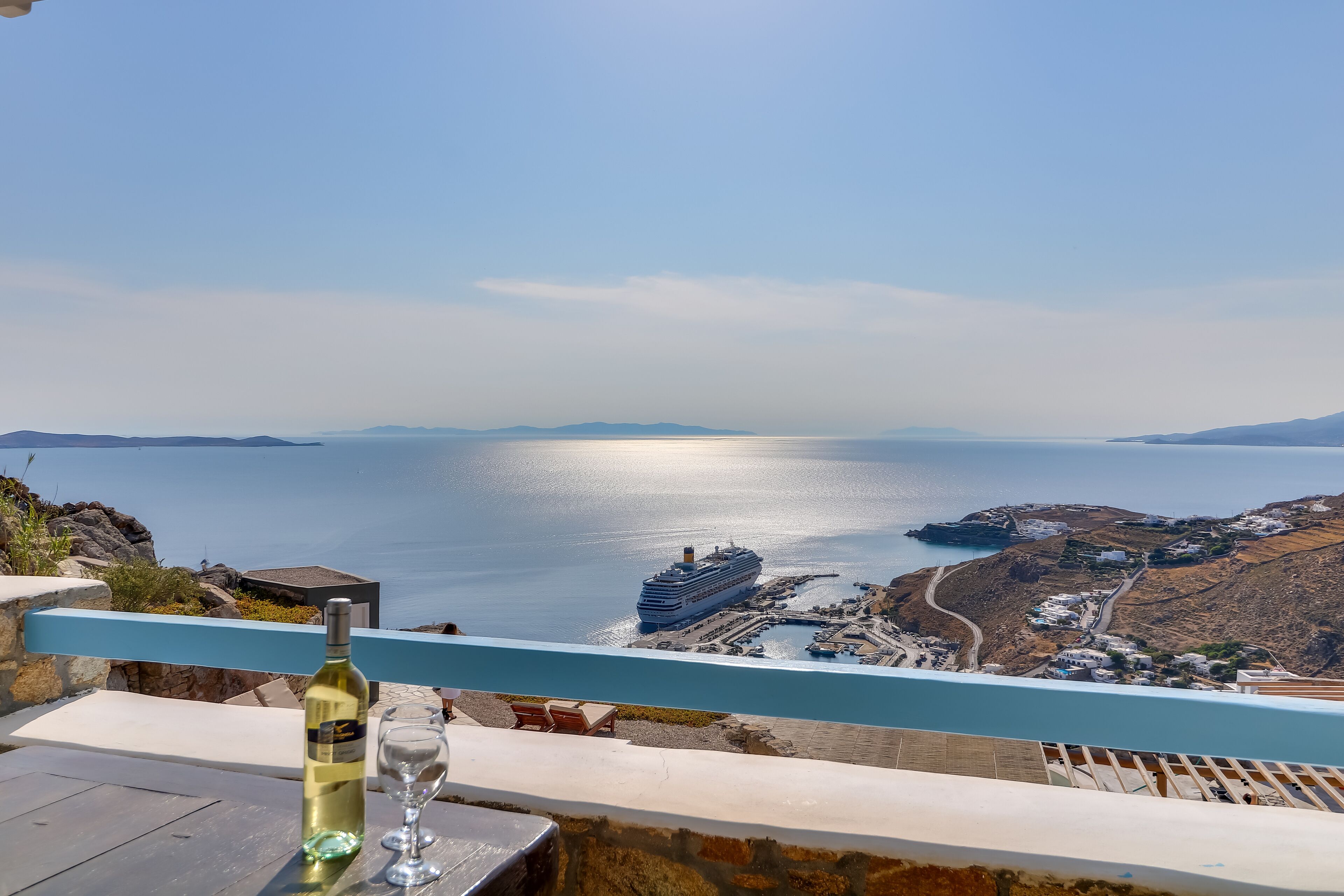 A scenic view from a balcony overlooking a coastal area with a cruise ship, a clear sky, and a bottle of wine on a table.