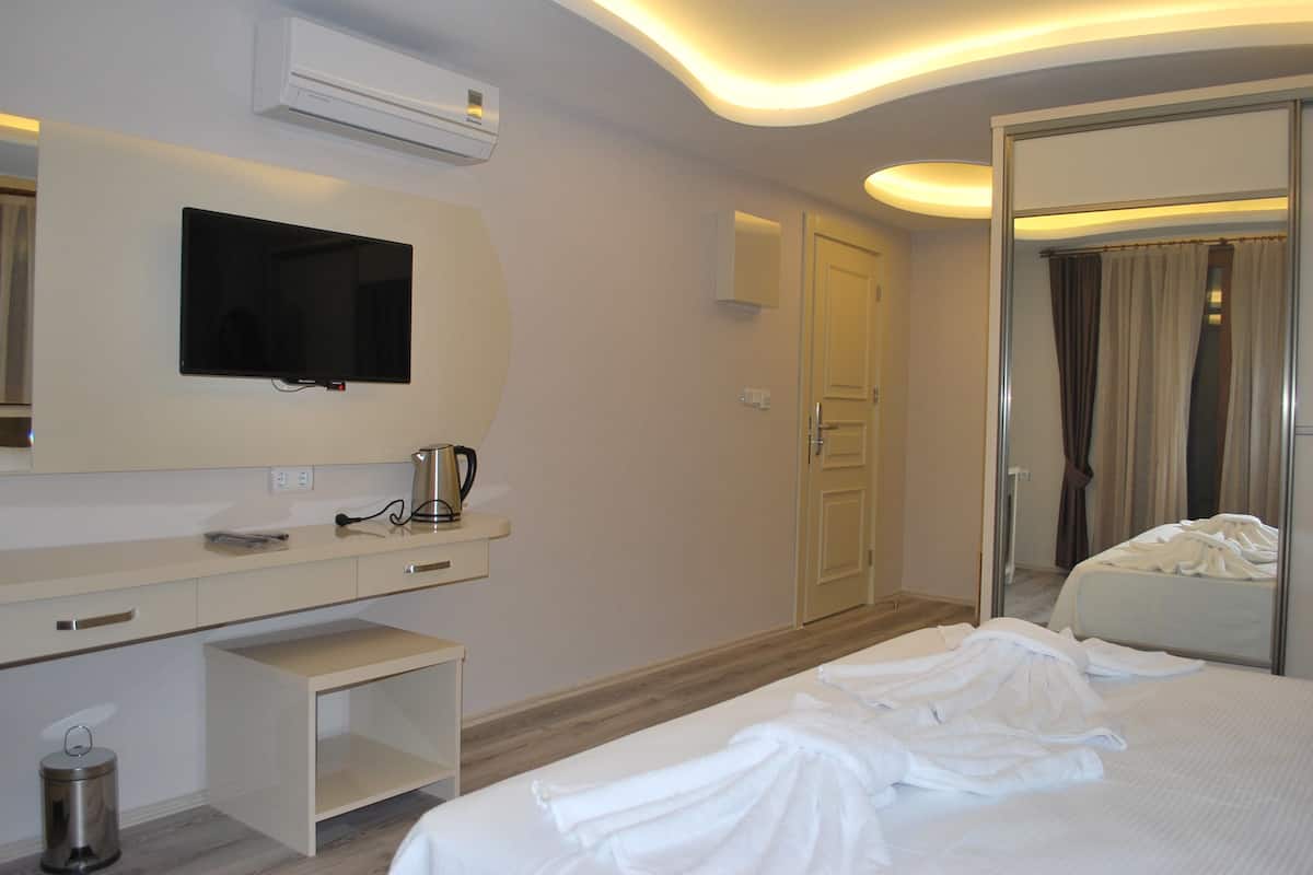 A hotel room with a bed, a TV mounted on the wall, a bedside table with a kettle, a trash bin, and a mirror reflecting the room.