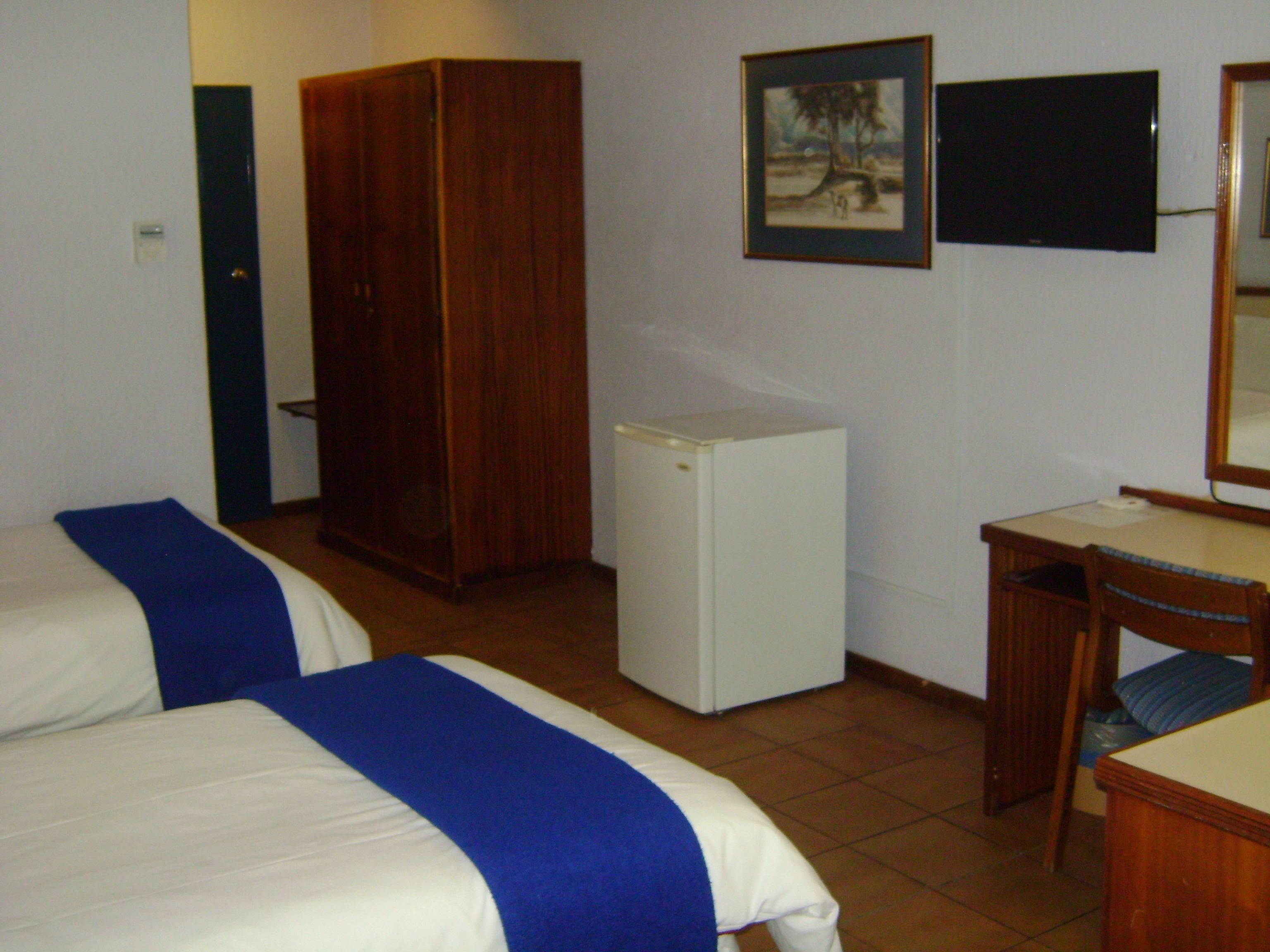 A bedroom with two beds, a small white refrigerator, a wooden wardrobe, a desk with a chair, and a framed picture on the wall.