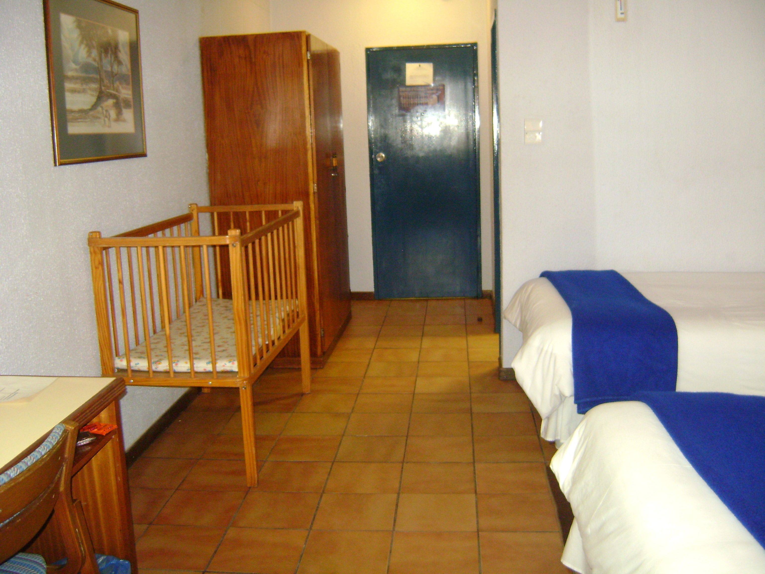 A room with a crib, a bed with a blue blanket, a wooden wardrobe, and a door with a notice.