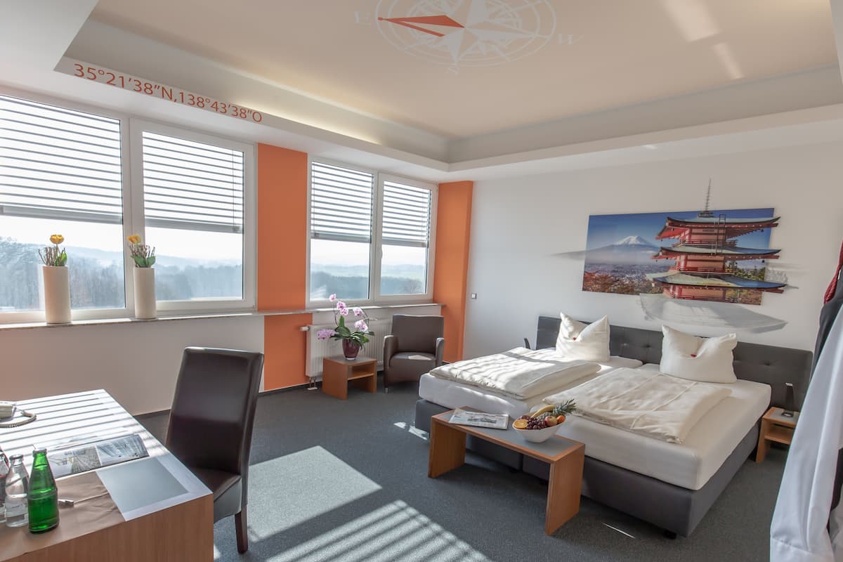 Habitación de hotel moderna con cama, escritorio y silla. Cuenta con ventanas con persianas, un cuadro de una pagoda y vistas a la montaña.
