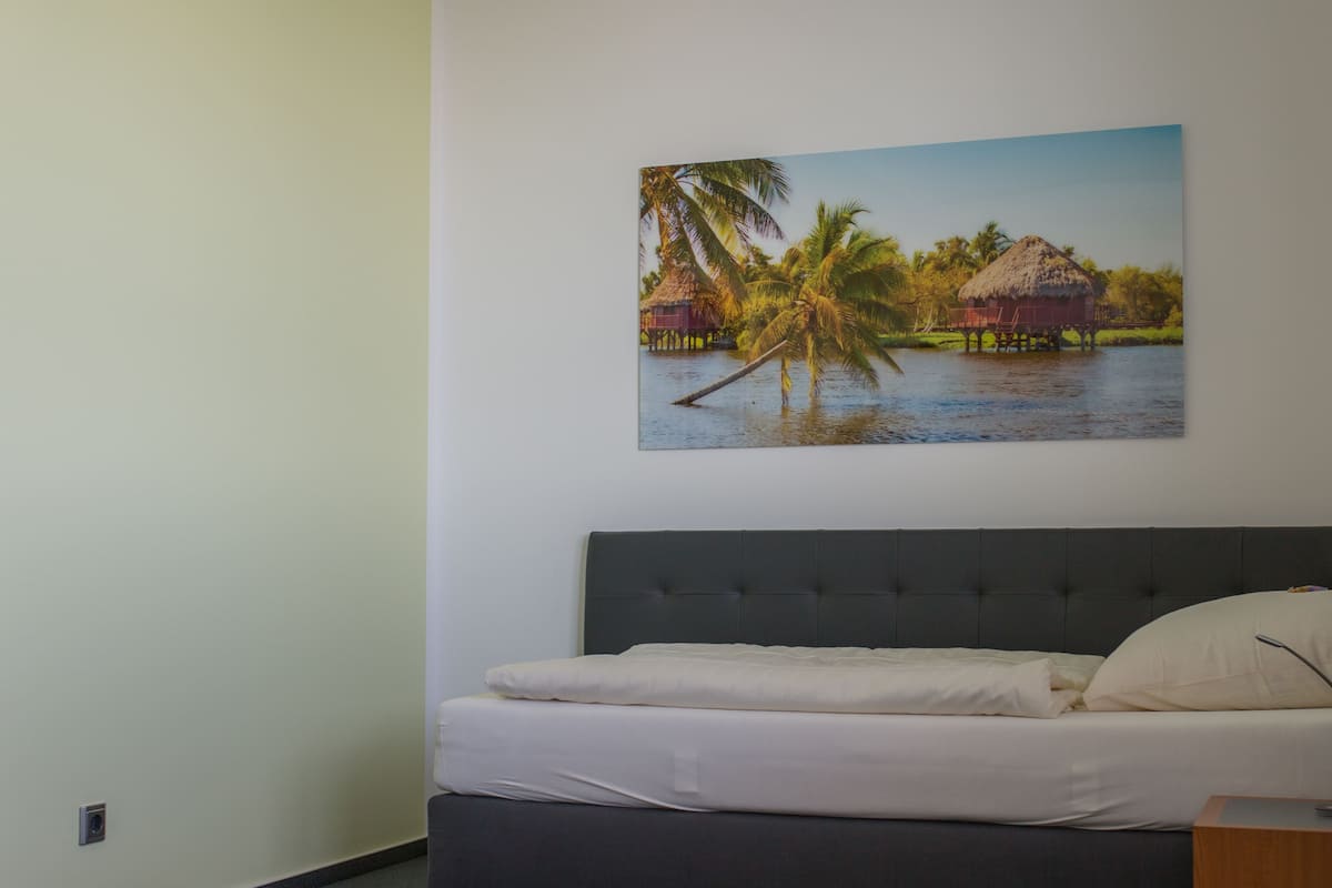 Un dormitorio con una cama, cabecera y un cuadro colgado en la pared de una escena tropical.