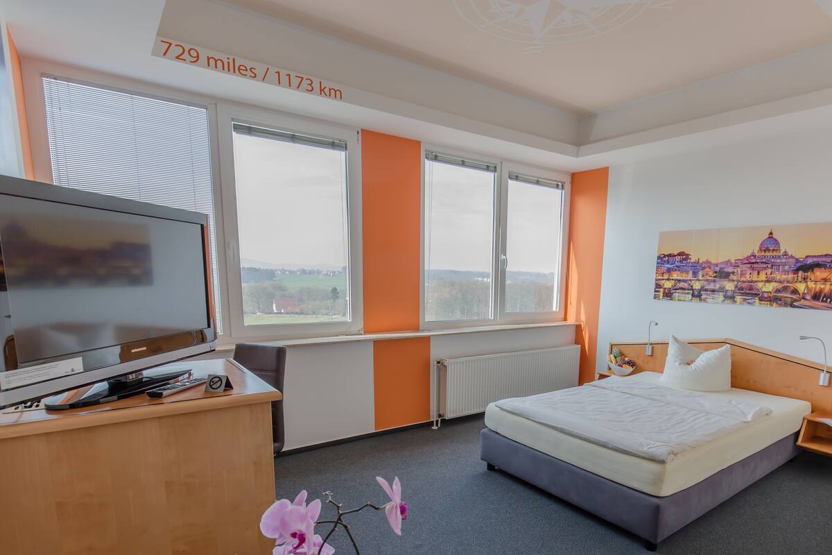 Habitación de hotel con cama, televisión y ventanas amplias con vista a la ciudad.