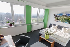 A hotel room with a bed, a desk, a chair, a window with a view of the countryside, and a painting on the wall.