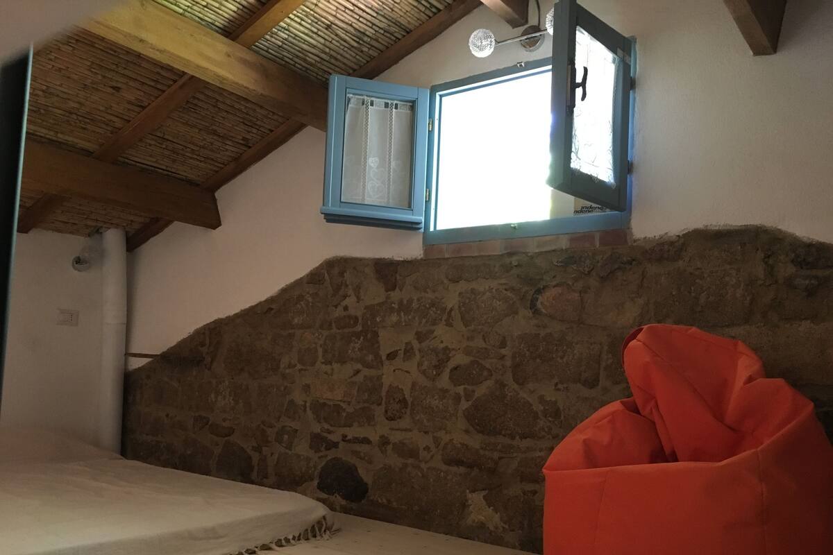 A room with a stone wall, a wooden ceiling, a bed, a red bean bag, and a window with a blue frame.