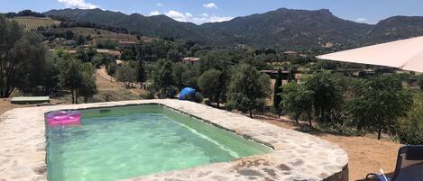 Una piscina de piedra con agua cristalina, rodeada por un muro de piedra, con vistas a un paisaje montañoso con árboles y casas.
