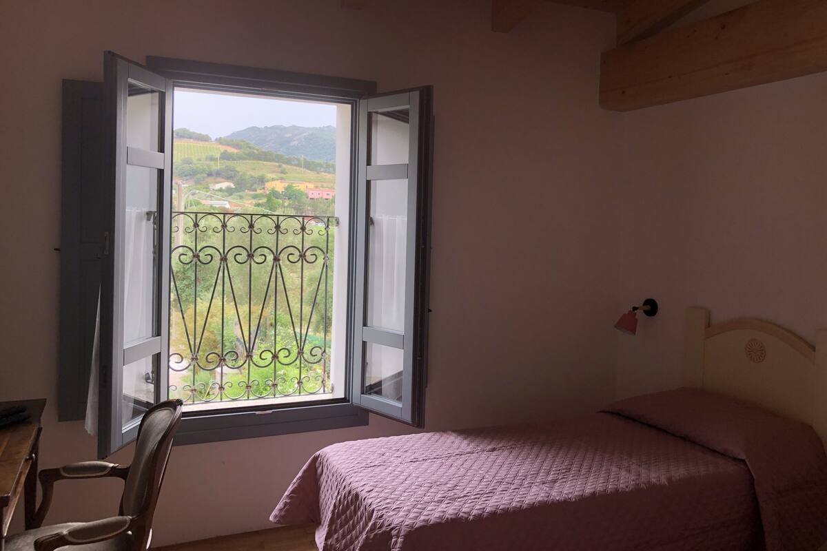 A bedroom with a bed, a chair, a window with a view of greenery and hills, and a wooden ceiling.