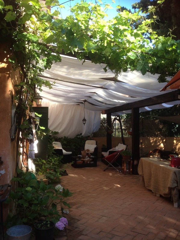 A covered patio with a table and chairs, surrounded by potted plants and a pergola with vines.