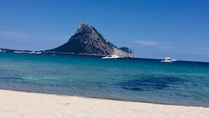 A beach with clear turquoise water, a sandy shore, and a prominent rocky mountain in the background. - La Casa delle Fate (Loiri Porto San Paolo)