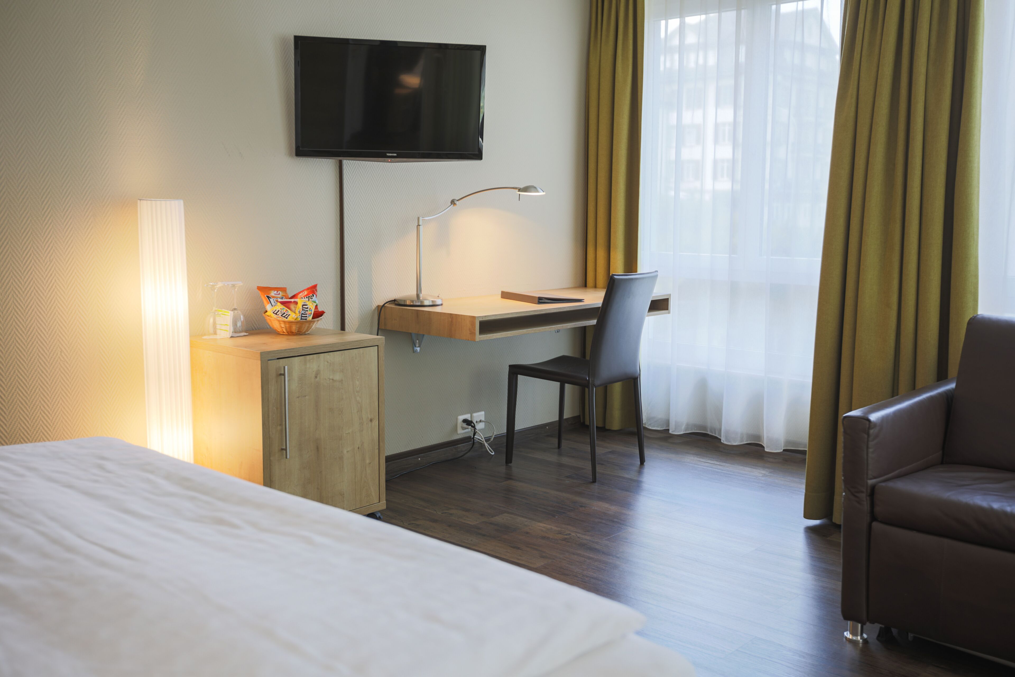 Habitación de hotel con cama, escritorio, silla, televisor y ventana con cortinas.