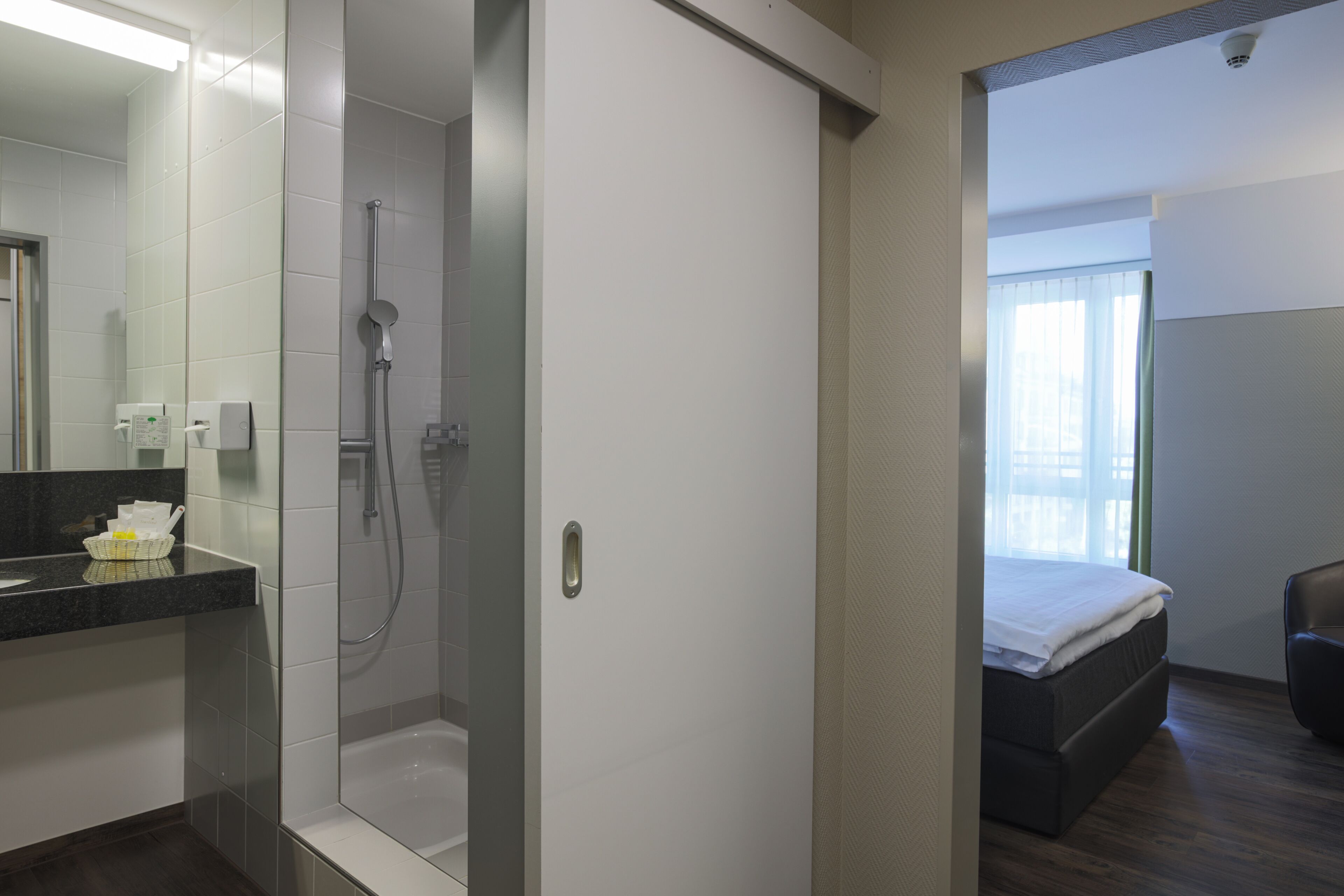 A modern bathroom with a shower, a toilet, and a sink. There is a bed with a white mattress and a brown headboard in the adjacent room.