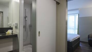 A modern bathroom with a shower, a toilet, and a sink. There is a bed with a white mattress and a brown headboard in the adjacent room.