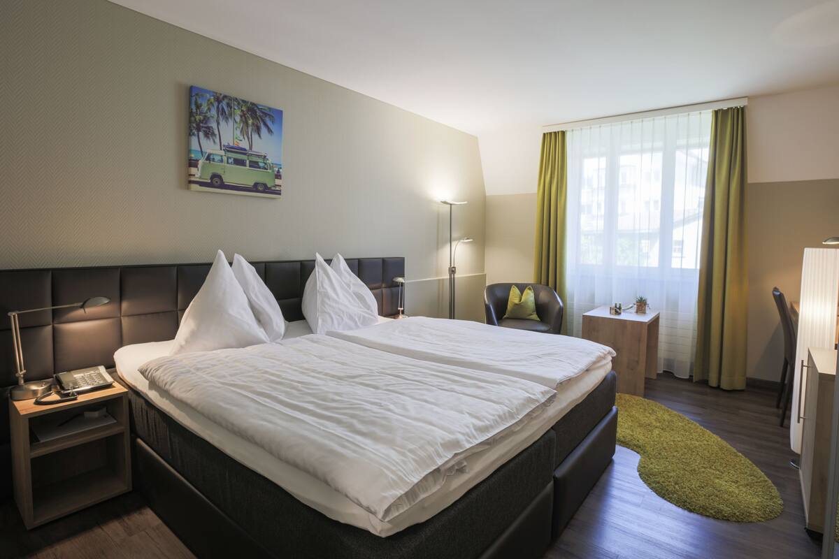 Una habitación de hotel con una cama grande, una mesita de noche con lámpara, una pequeña mesa con una planta y una ventana con cortinas.