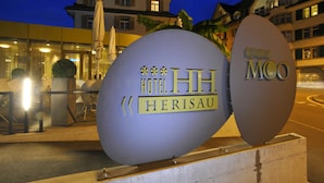 Een groot, verlicht bord met Hotel Herisau MCO, met een modern gebouw op de achtergrond.