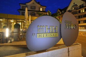 Un cartel grande e iluminado del Hotel Herisau MCO, con un edificio moderno al fondo.
