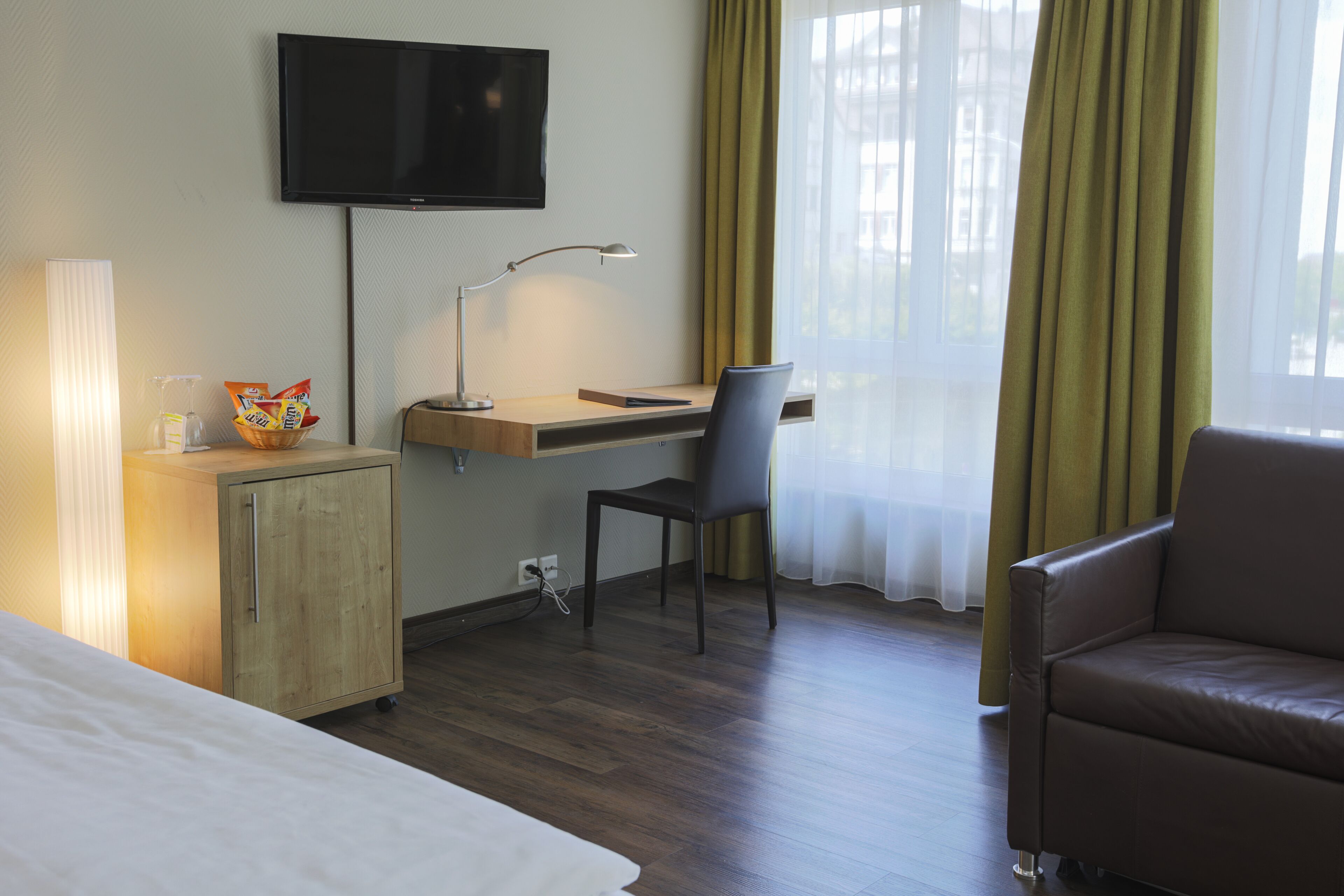 Une chambre d’hôtel comprenant un lit, un bureau avec une chaise, une télévision, une lampe, une armoire et une fenêtre avec des rideaux.