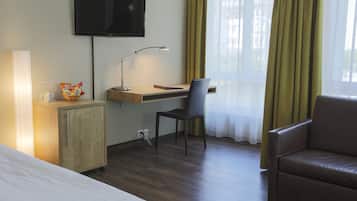 Une chambre d’hôtel comprenant un lit, un bureau avec une chaise, une télévision, une lampe, une armoire et une fenêtre avec des rideaux.