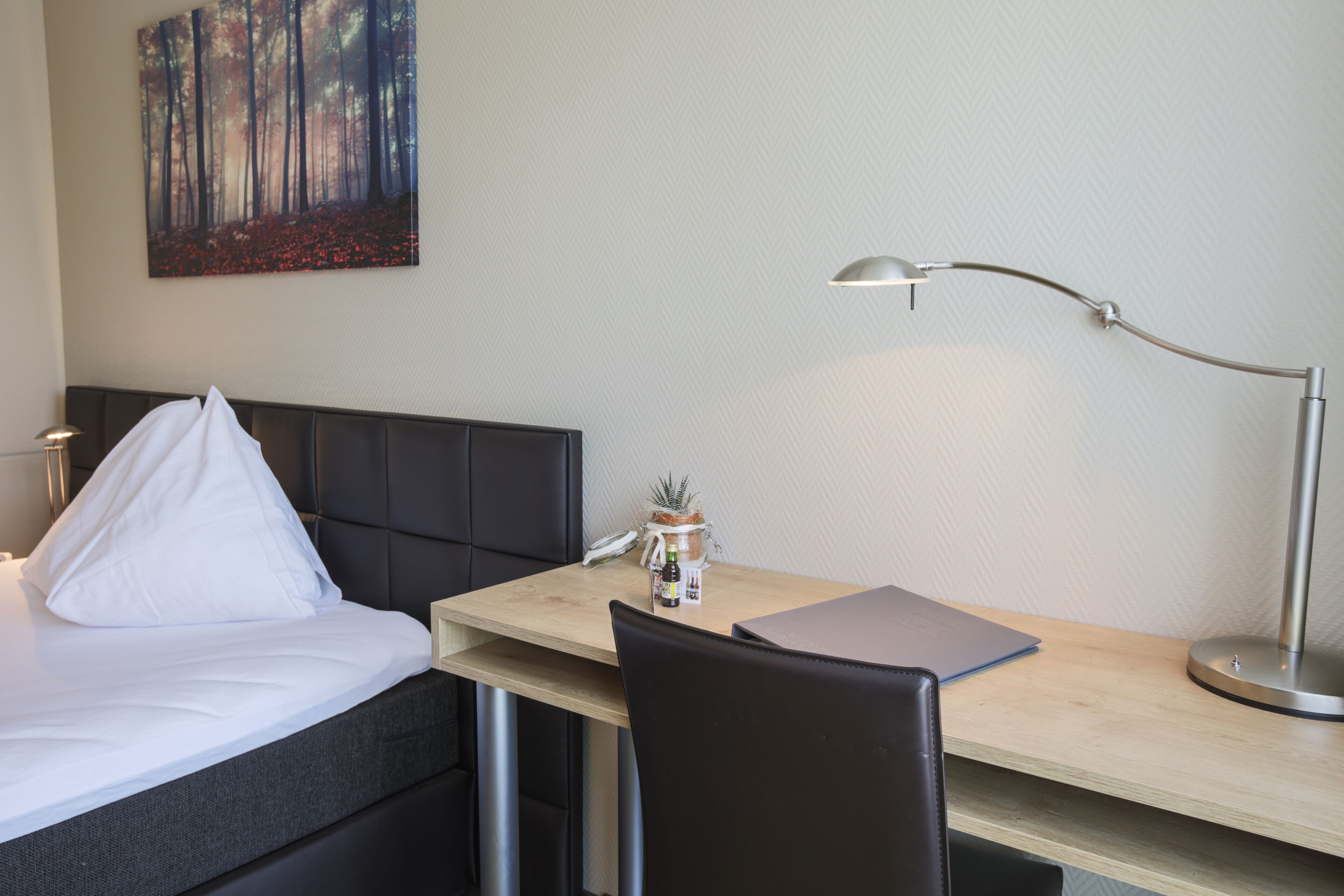 A hotel room with a bed, a desk, a chair, a lamp, a small plant, and a painting on the wall.