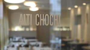 Een onscherp zicht in een restaurant met tafels en stoelen, en de naam "ALTI CHOCHI" zichtbaar op het raam.
