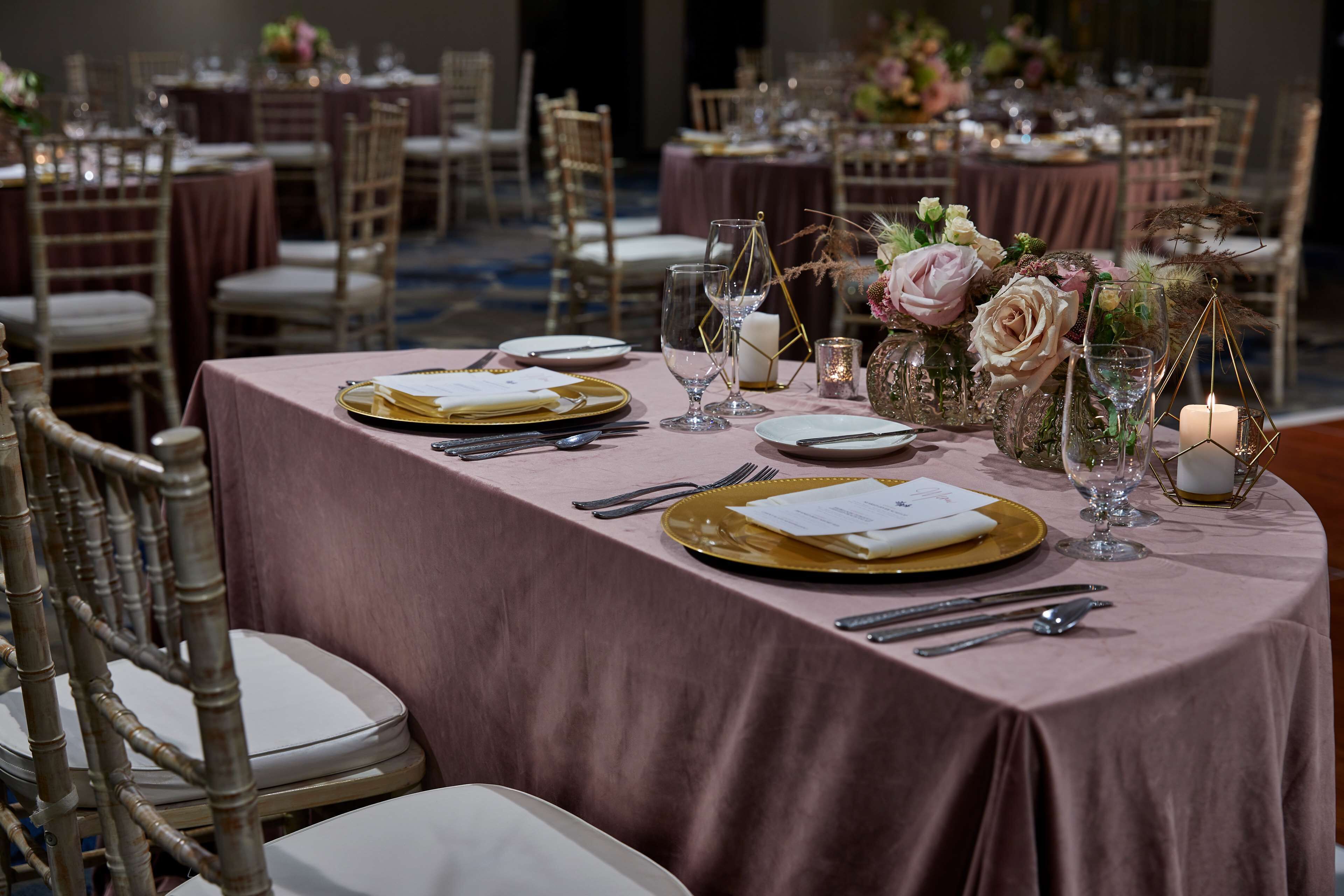 A banquet hall with round tables set for a formal event, featuring gold-rimmed plates, clear glassware, and floral centerpieces.