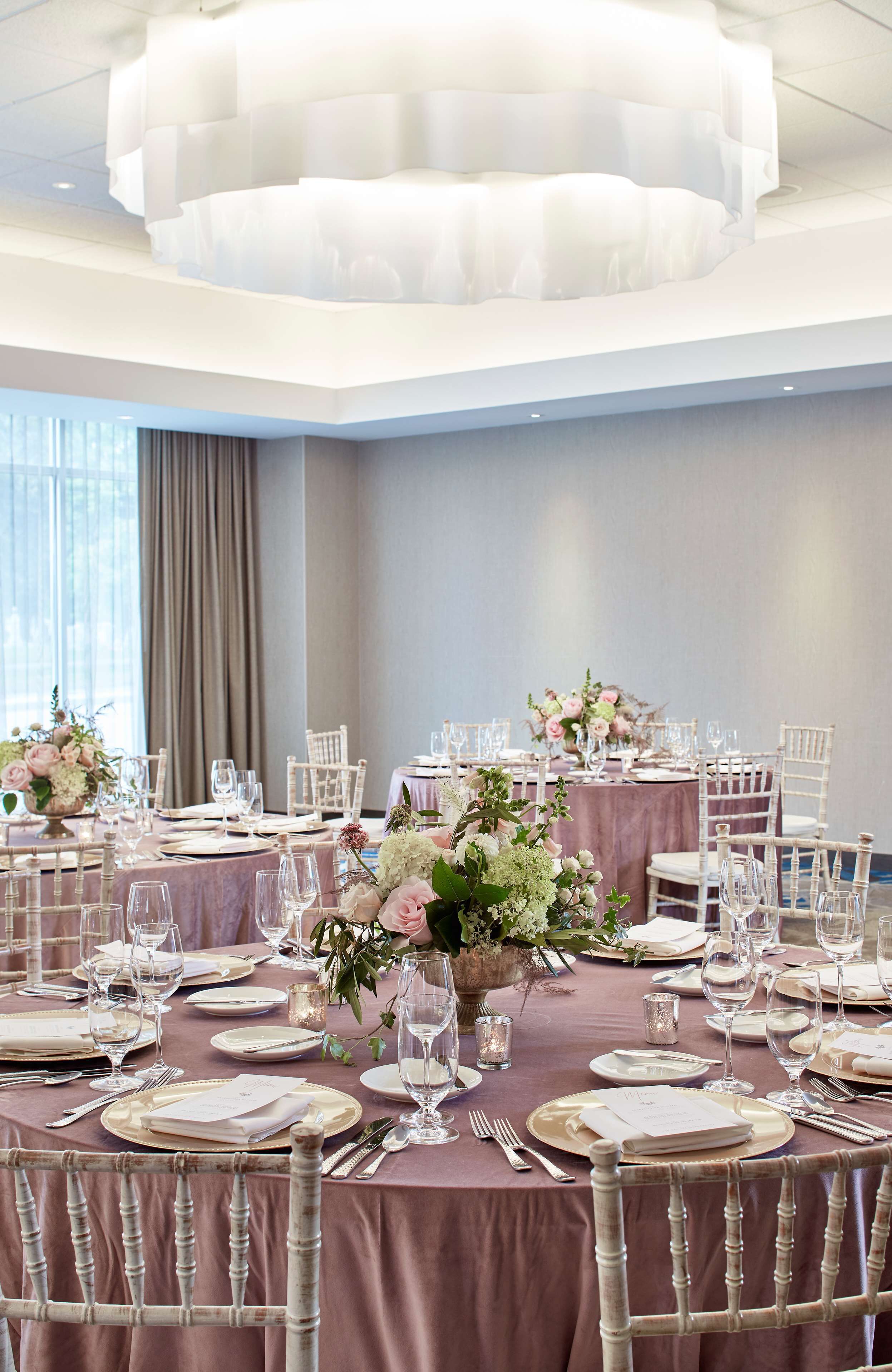 Salão de festas com mesas redondas montadas para um evento formal, com cadeiras brancas e elegantes arranjos de mesa.