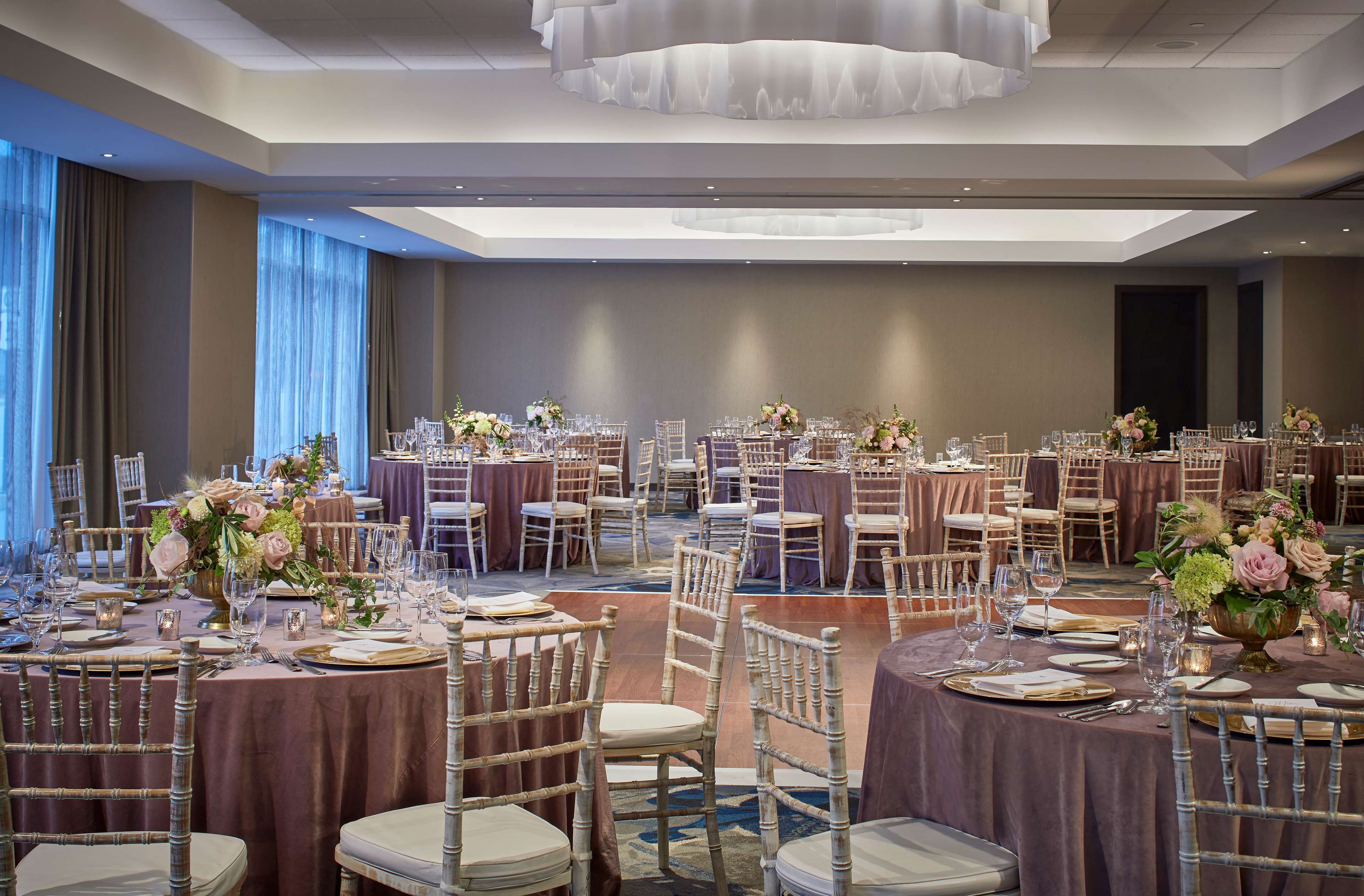 Een feestzaal met ronde tafels, stoelen en bloemstukken als decoratie.