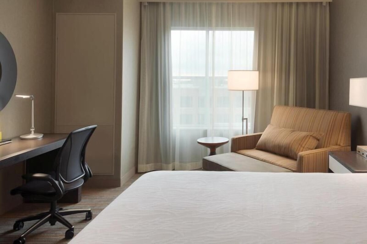A hotel room with a bed, a desk with a chair, a small table, and a lamp.