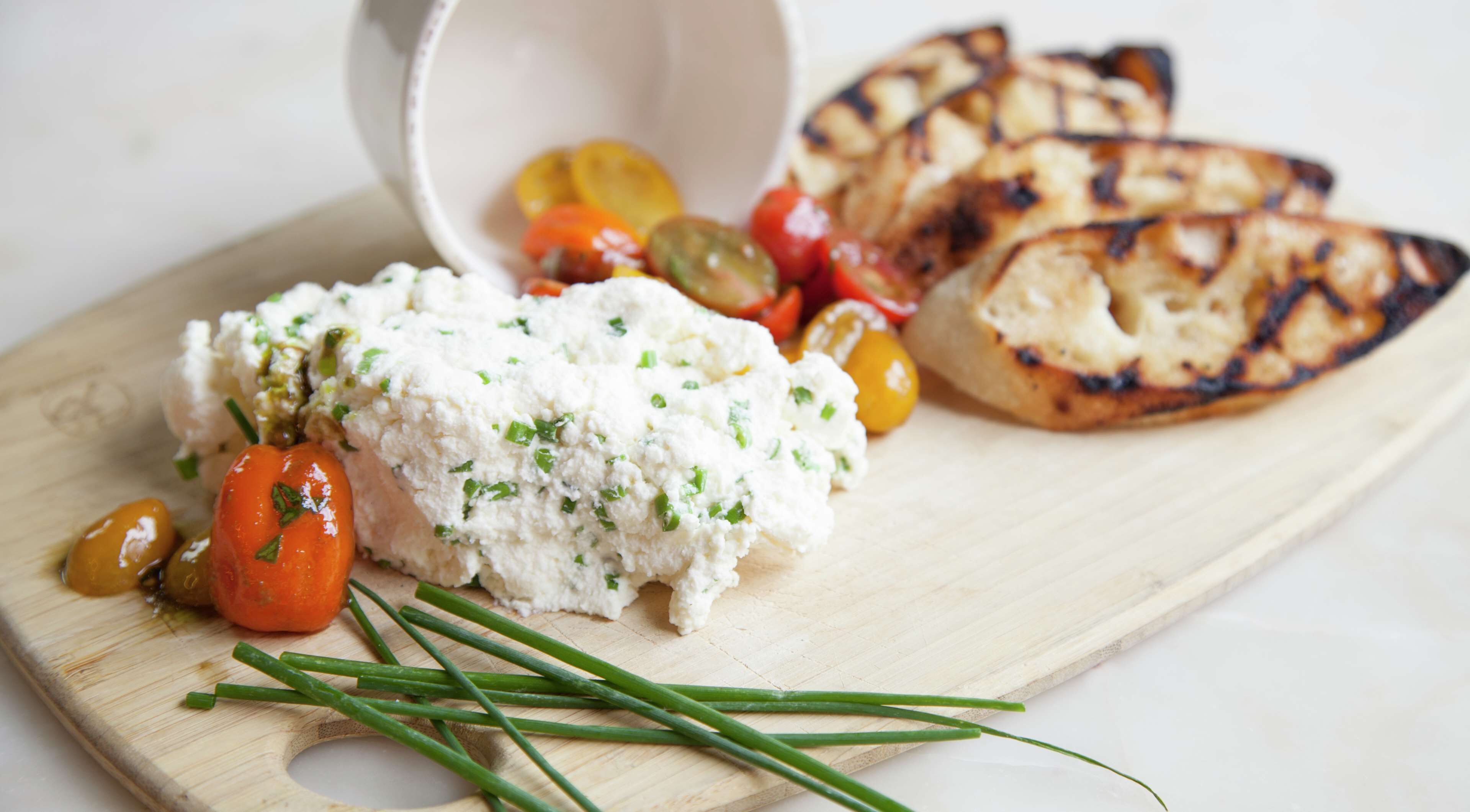 Una tabla de madera con queso blanco adornado con hierbas, verduras a la parrilla y rebanadas de pan.
