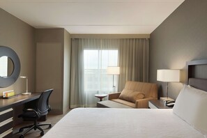 A hotel room with a large bed, a desk with a chair, a sofa, a round mirror, and a window with curtains.