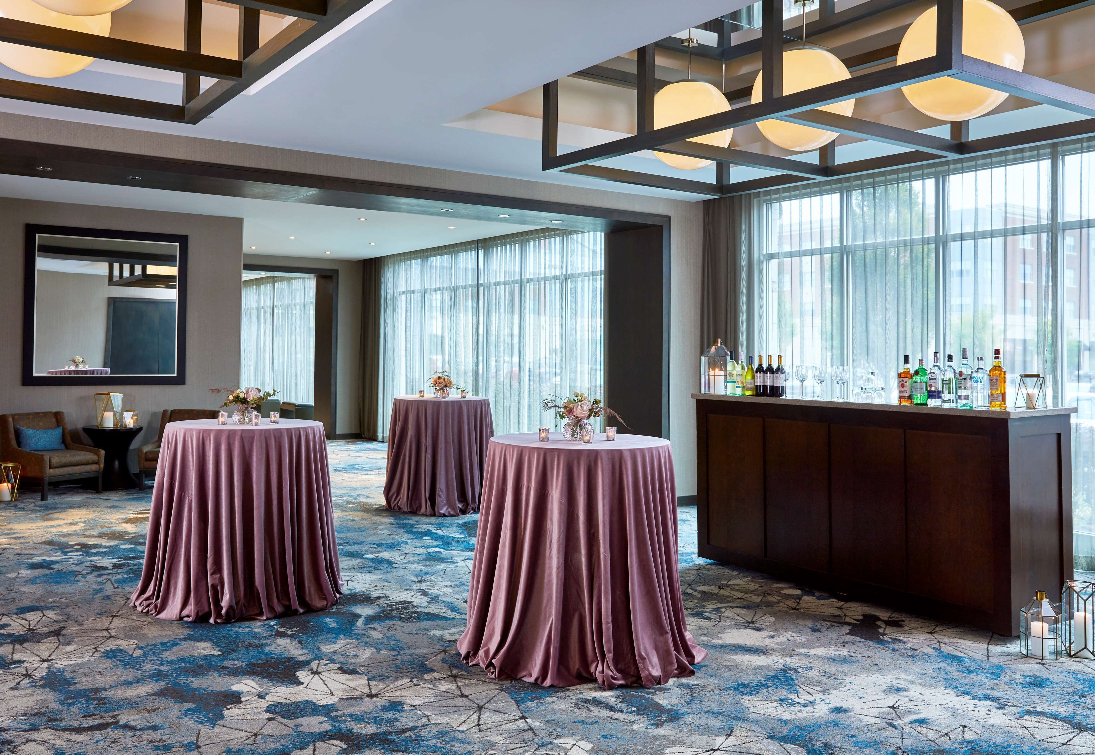 A hotel lobby with three round tables covered in purple tablecloths, a bar area with bottles, and large windows allowing natural light.