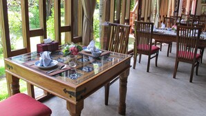 A dining area with a glass-top table, wooden chairs, and a view of greenery outside. - Dhabkwan Resort (Nonthaburi)