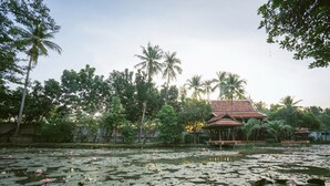 A serene pond with lily pads and lotus flowers, surrounded by lush greenery and a traditional building with a tiled roof. - Dhabkwan Resort (Nonthaburi)