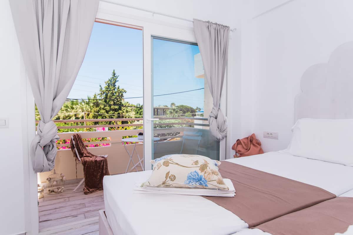 Ein Hotelzimmer mit Bett, Stuhl und Blick auf einen Balkon mit Pflanzen und einem Baum.