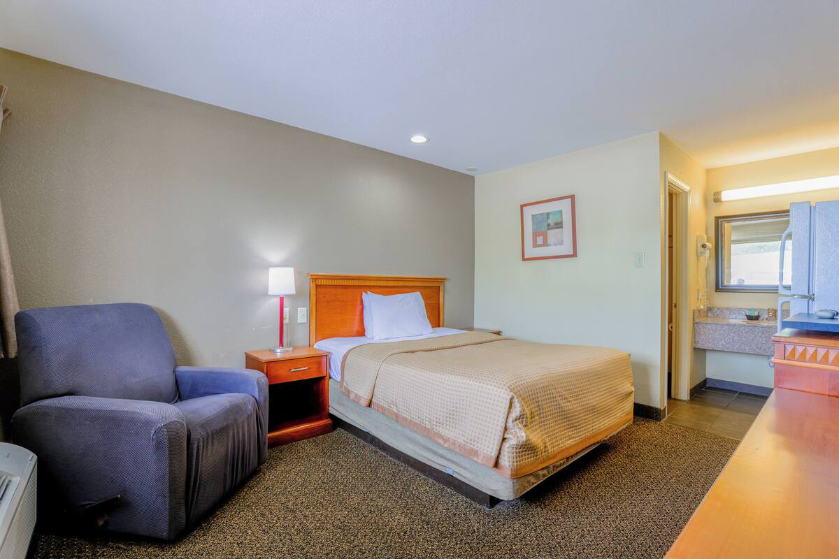 A hotel room with a bed, a chair, a nightstand, a desk, and a bathroom visible through an open door.