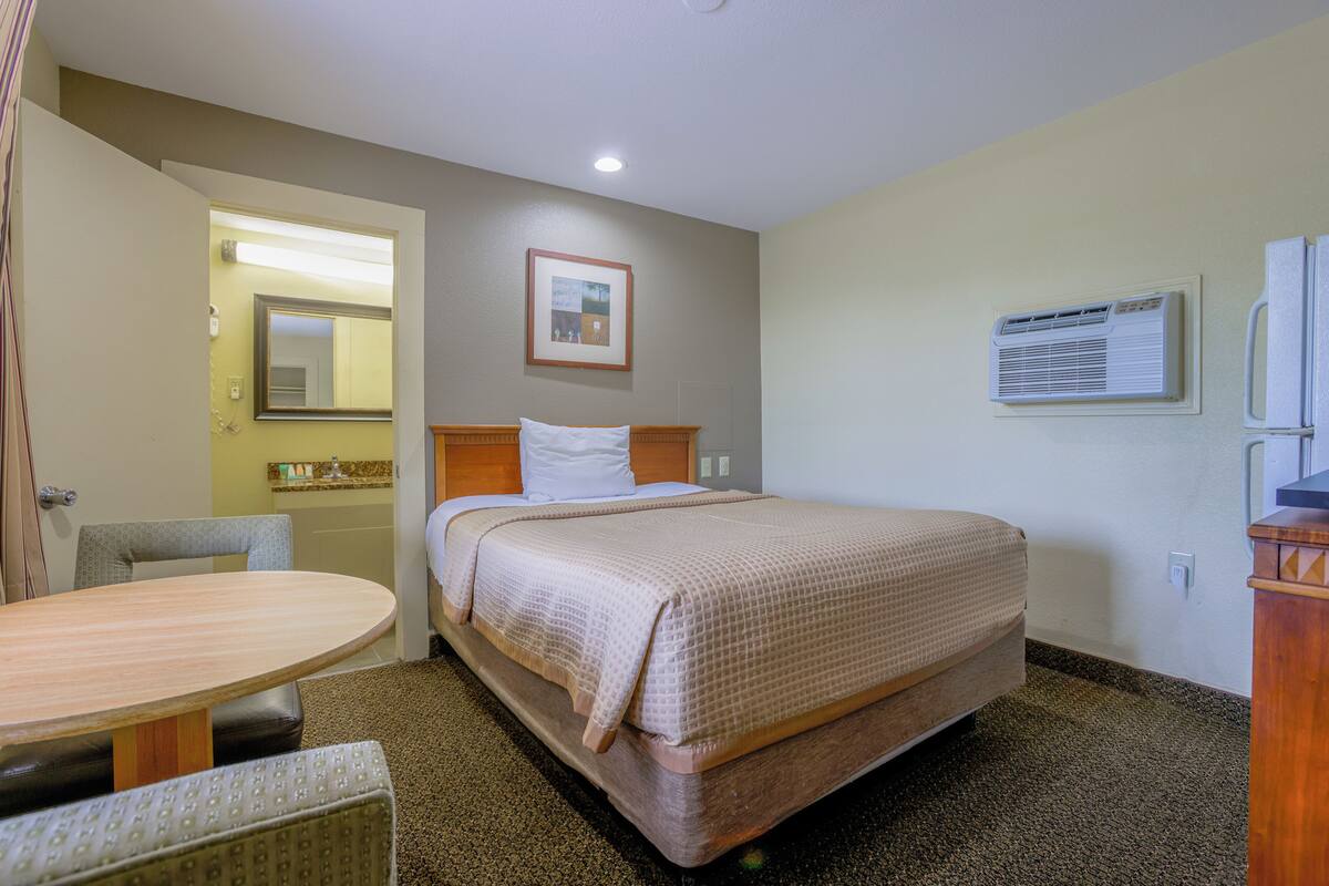 A hotel room with a bed, a bedside table, a chair, a small table, a mirror, and a picture on the wall.