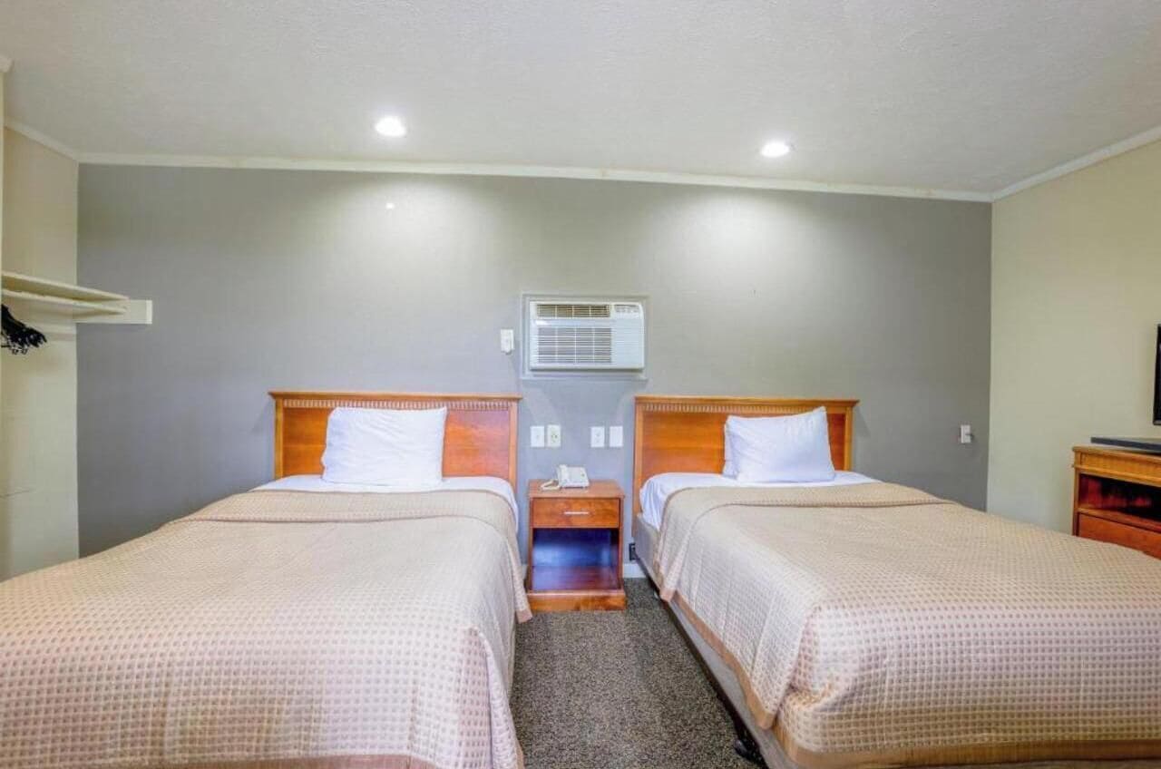 A hotel room with two single beds, a small bedside table with a lamp, an air conditioning unit on the wall, and a TV stand with a TV.