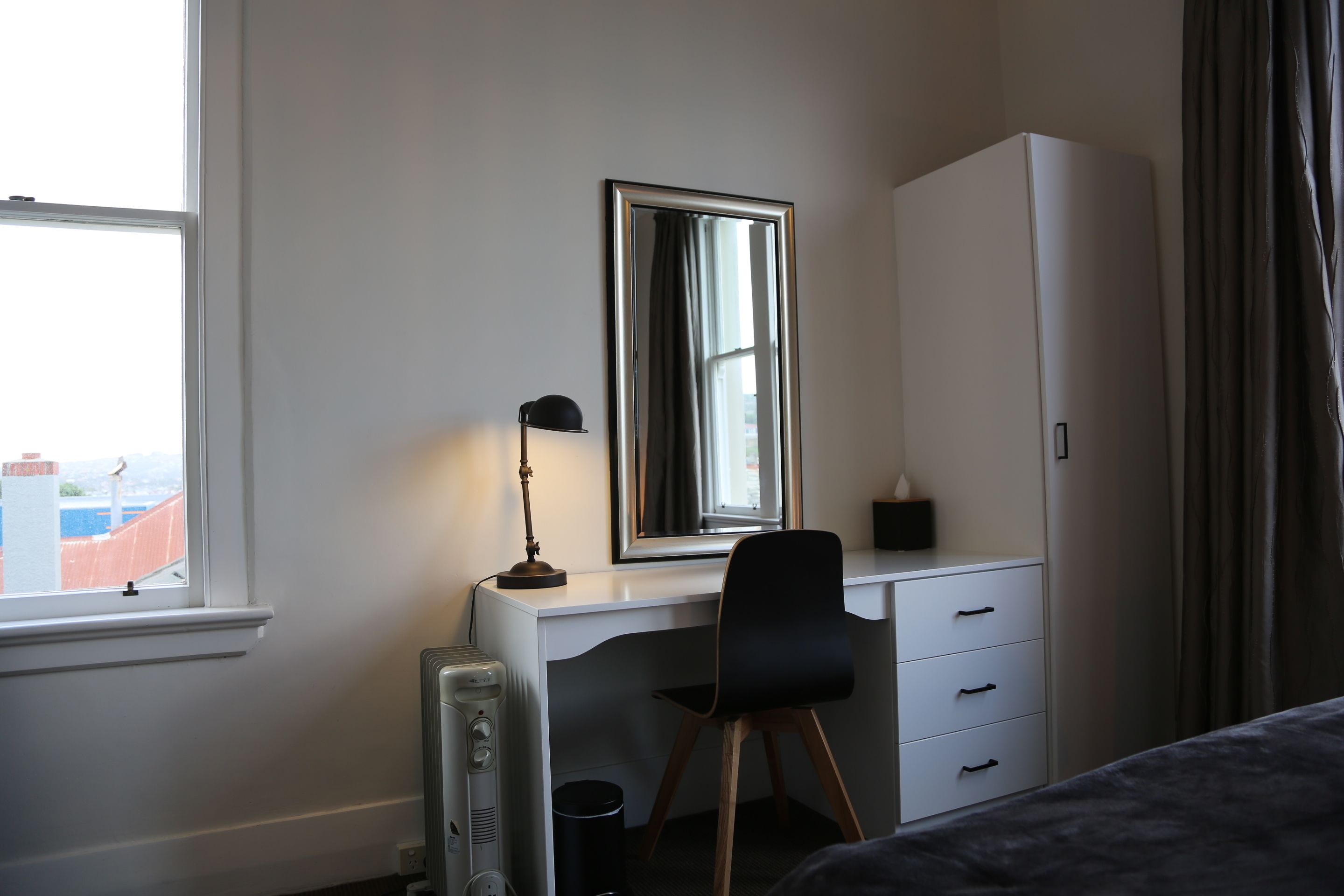 A bedroom with a bed, a desk with a chair, a radiator, a mirror, and a wardrobe.
