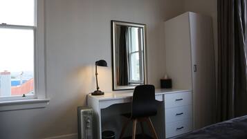 A bedroom with a bed, a desk with a chair, a radiator, a mirror, and a wardrobe.