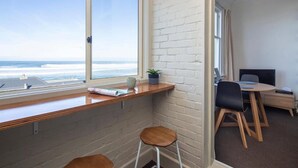 A corner room with a large window overlooking the ocean, a desk with a chair, and a small stool. - Majestic Mansions - Apartments at St Clair (Dunedin)