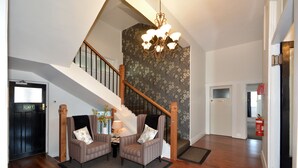 A spacious living area with a wooden staircase, two armchairs, a table with a lamp, and a chandelier. - Majestic Mansions - Apartments at St Clair (Dunedin)