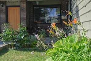 Un edificio de ladrillo con una puerta de madera, una ventana con un letrero y un jardín con diversas plantas.