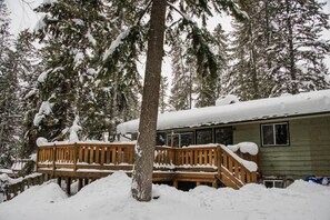 Eine Holzhütte mit einer Teakholzterrasse und einem mit Schnee bedeckten Dach, umgeben von immergrünen Bäumen.