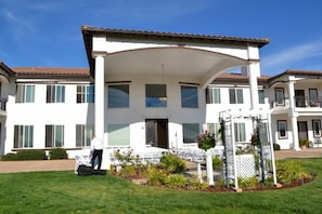 A large, two-story building with a covered patio area, multiple windows, and a well-maintained lawn. - Que Syrah (Temecula)