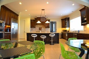 A modern kitchen with a central island, bar stools, and a dining area with green chairs. - Que Syrah (Temecula)
